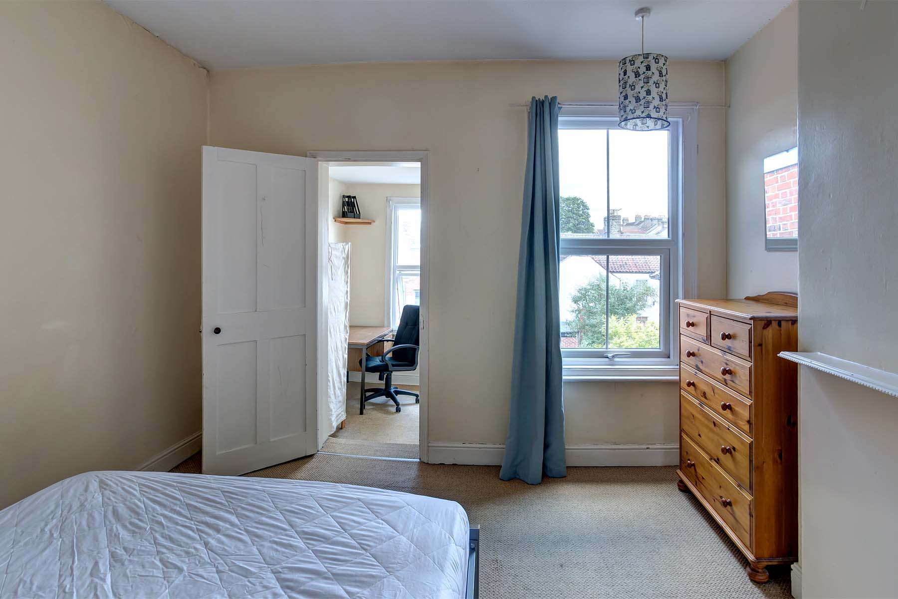 Double bedroom with wooden chest of drawers that joins a study room with a desk and a chair.