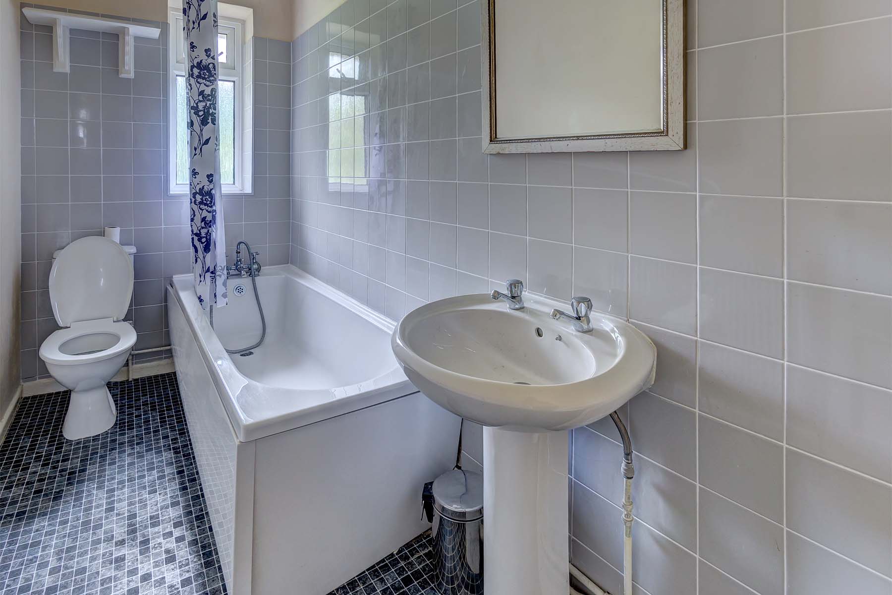 Tiled bathroom with white bath tub with shower over the bath. White toilet and sink, mirror above the sink.