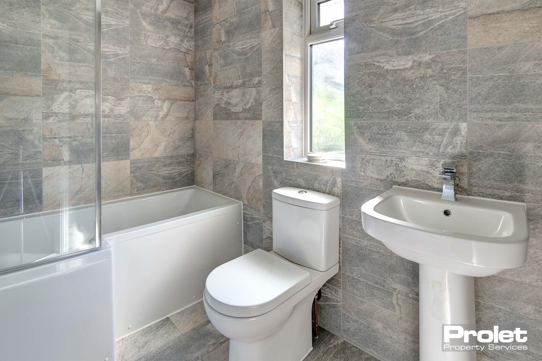 Stone coloured tiled bathroom with modern (white) sink, toilet and bath tub. Glass screen on the bath tub due to shower over the bath set up.