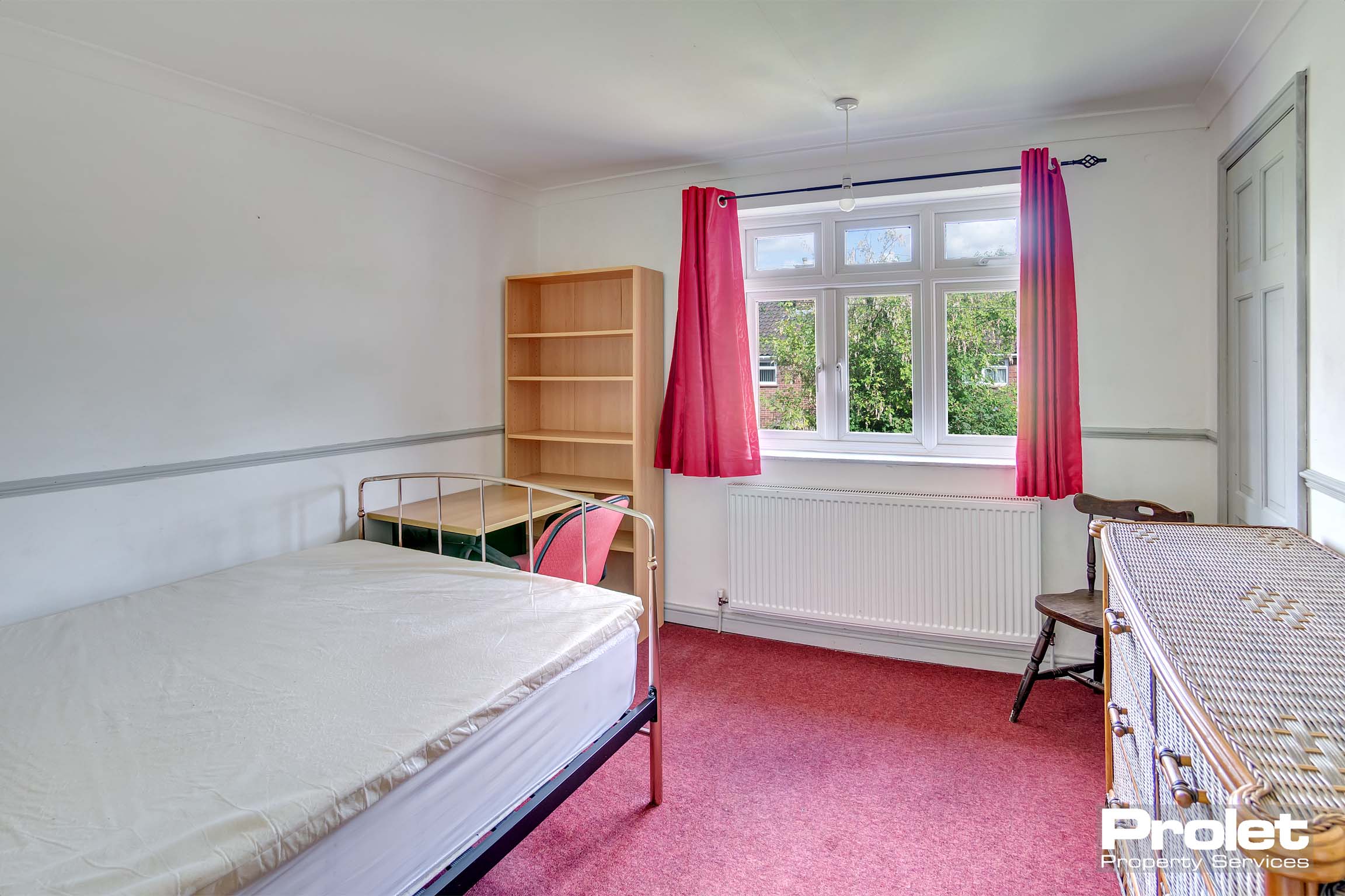 Double bedroom with red carpet and curtains, a wooden bookshelf, desk and chest of drawers.