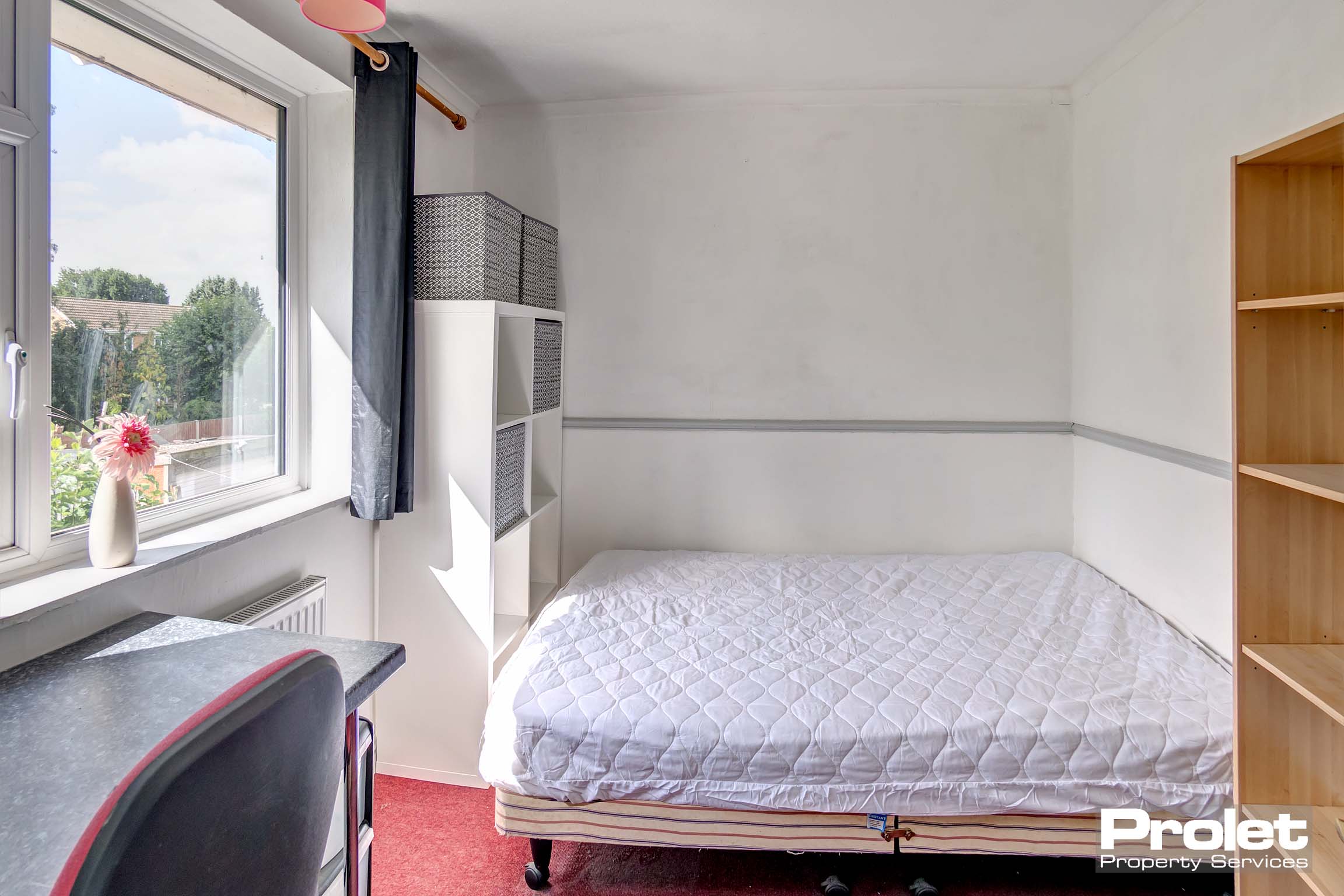 Double bedroom with red carpet and grey curtains. A coal coloured desk, a white storage unit and a wooden bookshelf.