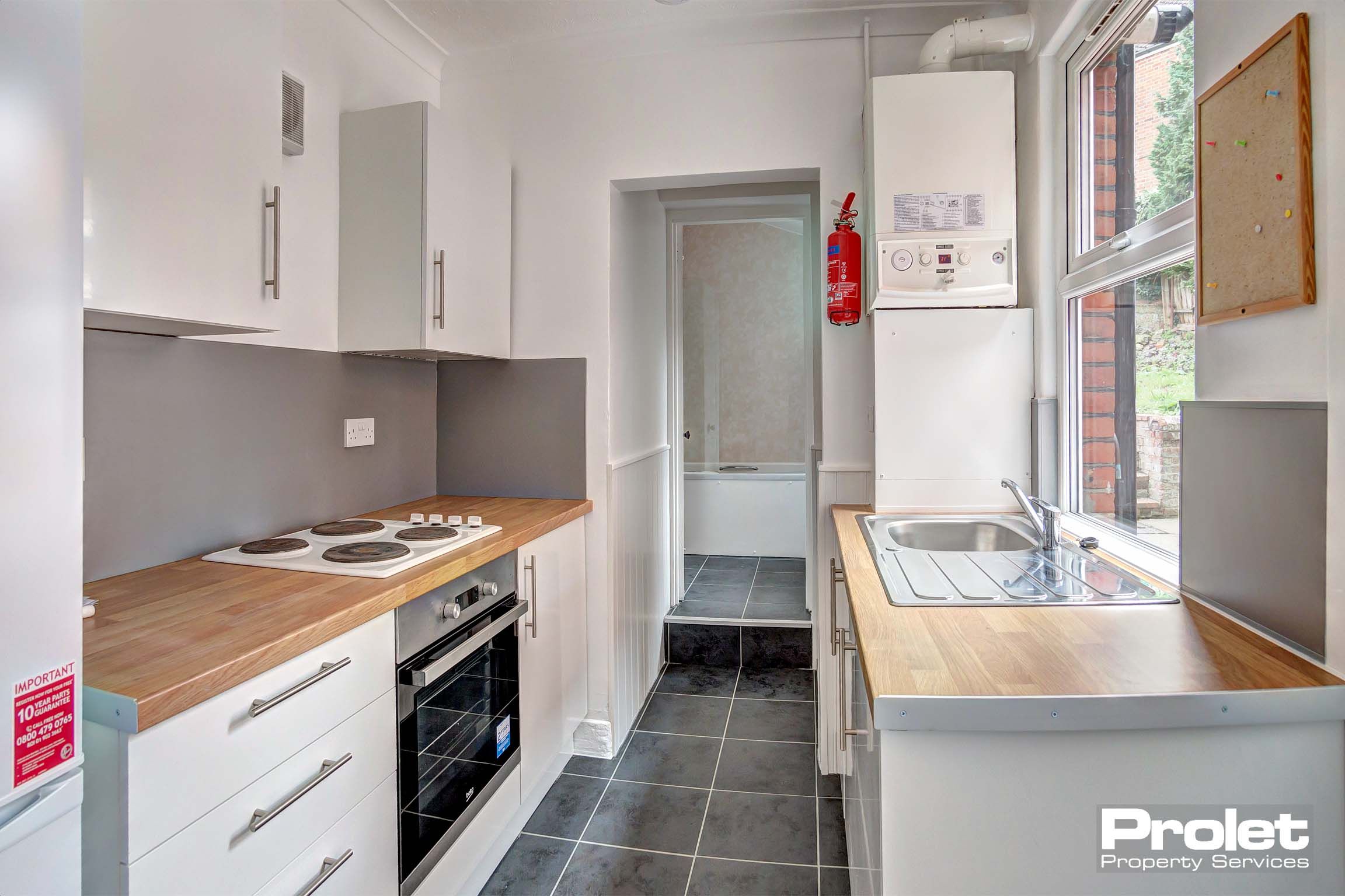 Galley style kitchen with white fitted kitchen units and a wooden worktop. Dark grey tiled flooring.