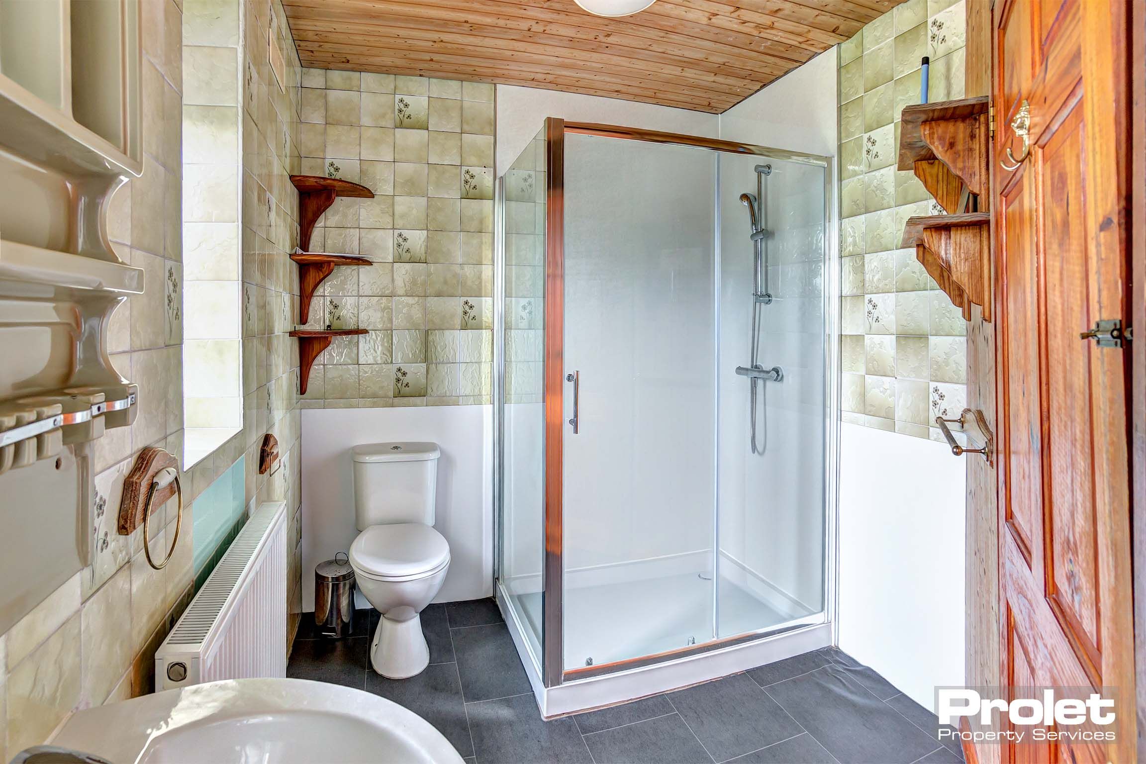 Bathroom with green tiled walls and dark grey flooring. A glass walk in shower and a white sink and toilet.