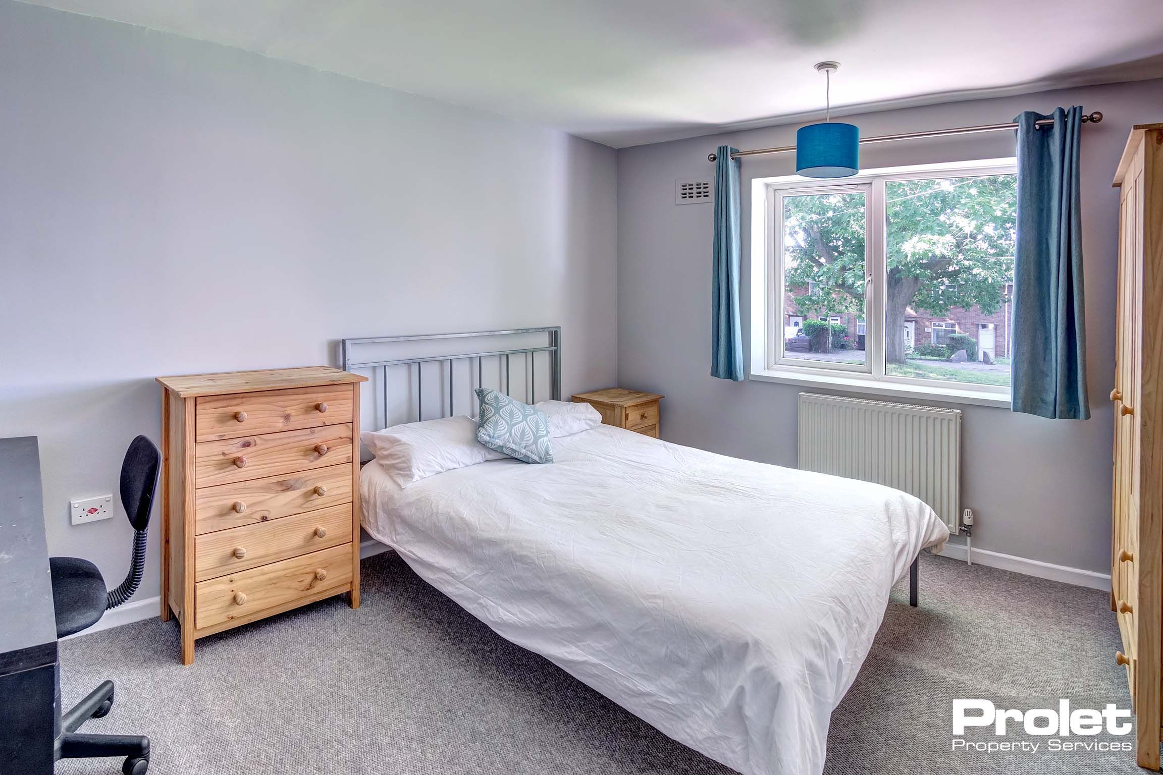 A double bedroom with a metal bed frame, a black desk and chair and a wooden chest of drawers, bedside table and wardrobe.