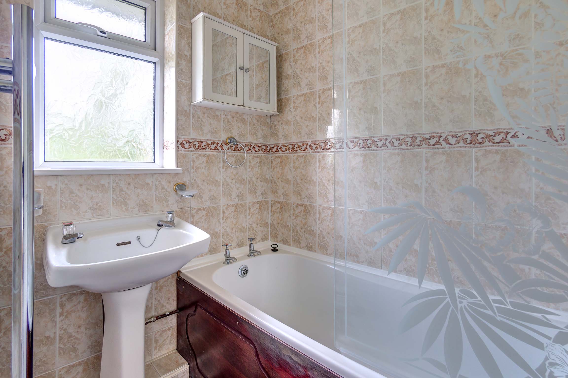 Bathroom with tiled walls and a bathtub with wooden bath panel. Shower over the bath with glass splash guard. White sink.