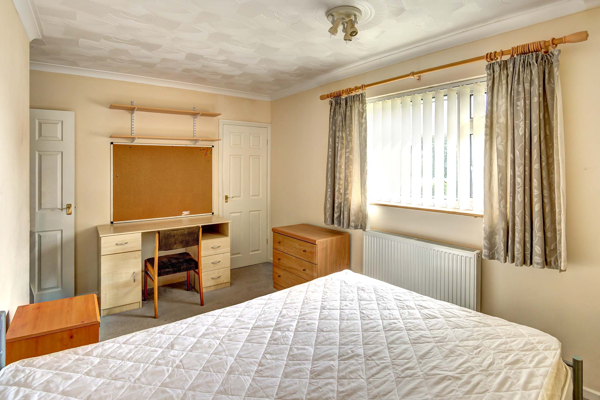 Large double bedroom with wooden bedside table and wooden desk and chair, with wooden cork board.