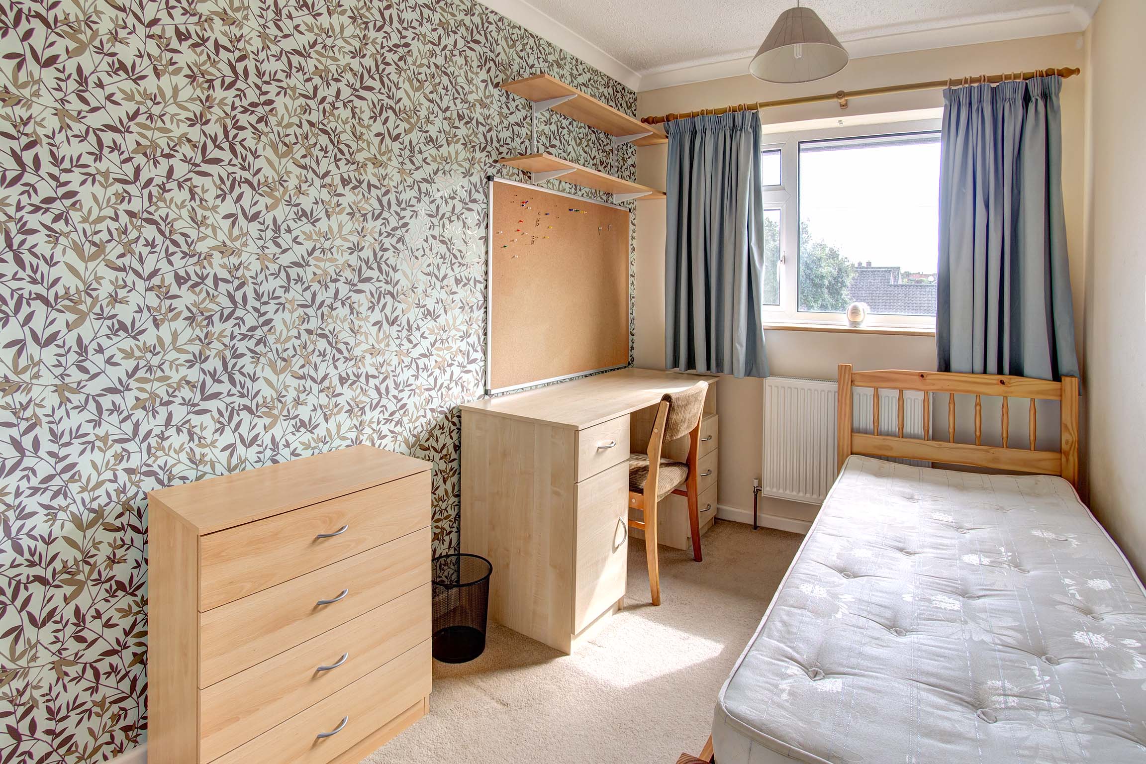 Bedroom with wooden bed frame, wallpaper on one wall. A wooden desk and chair and a set of chest of drawers.