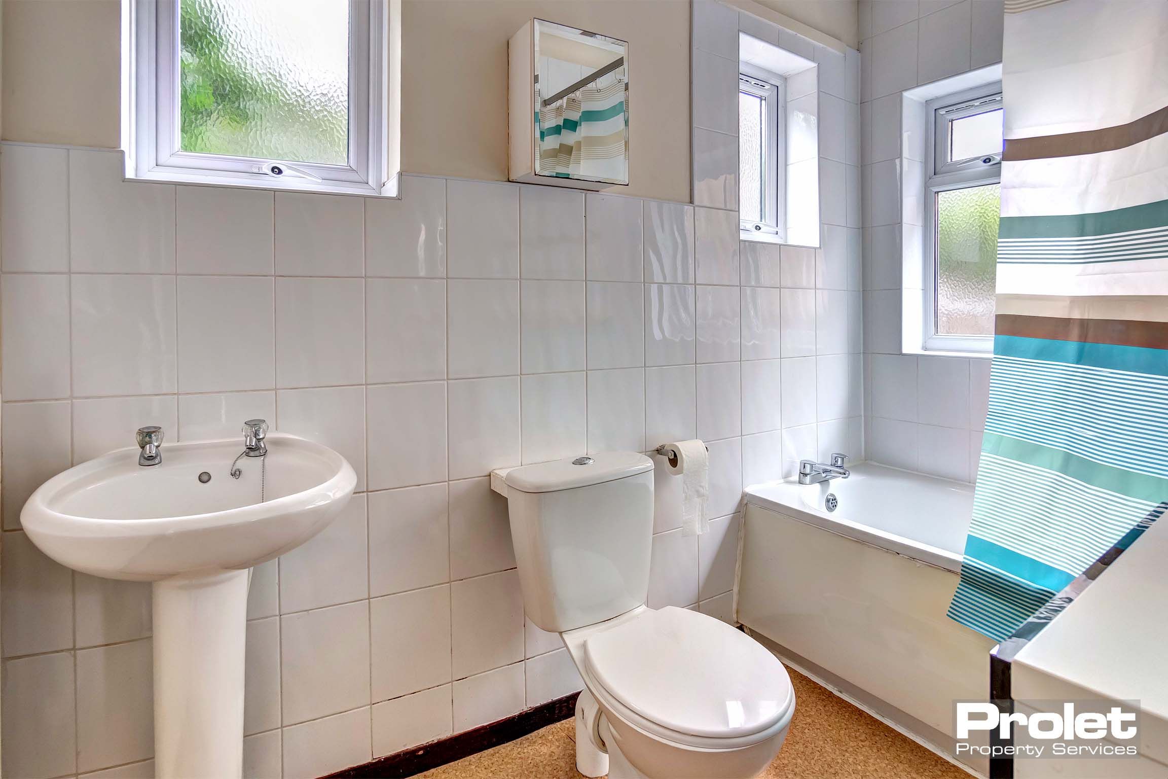 White tiled bathroom with white bath tub, toilet and sink with a shower over the bath.