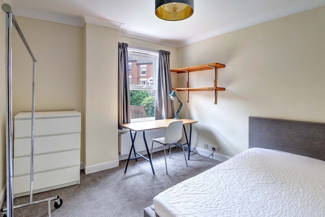 A double bedroom with a grey bedframe, shelves on the wall, a clothes rail, a white chest of drawers and a wooden desk with a white chair.