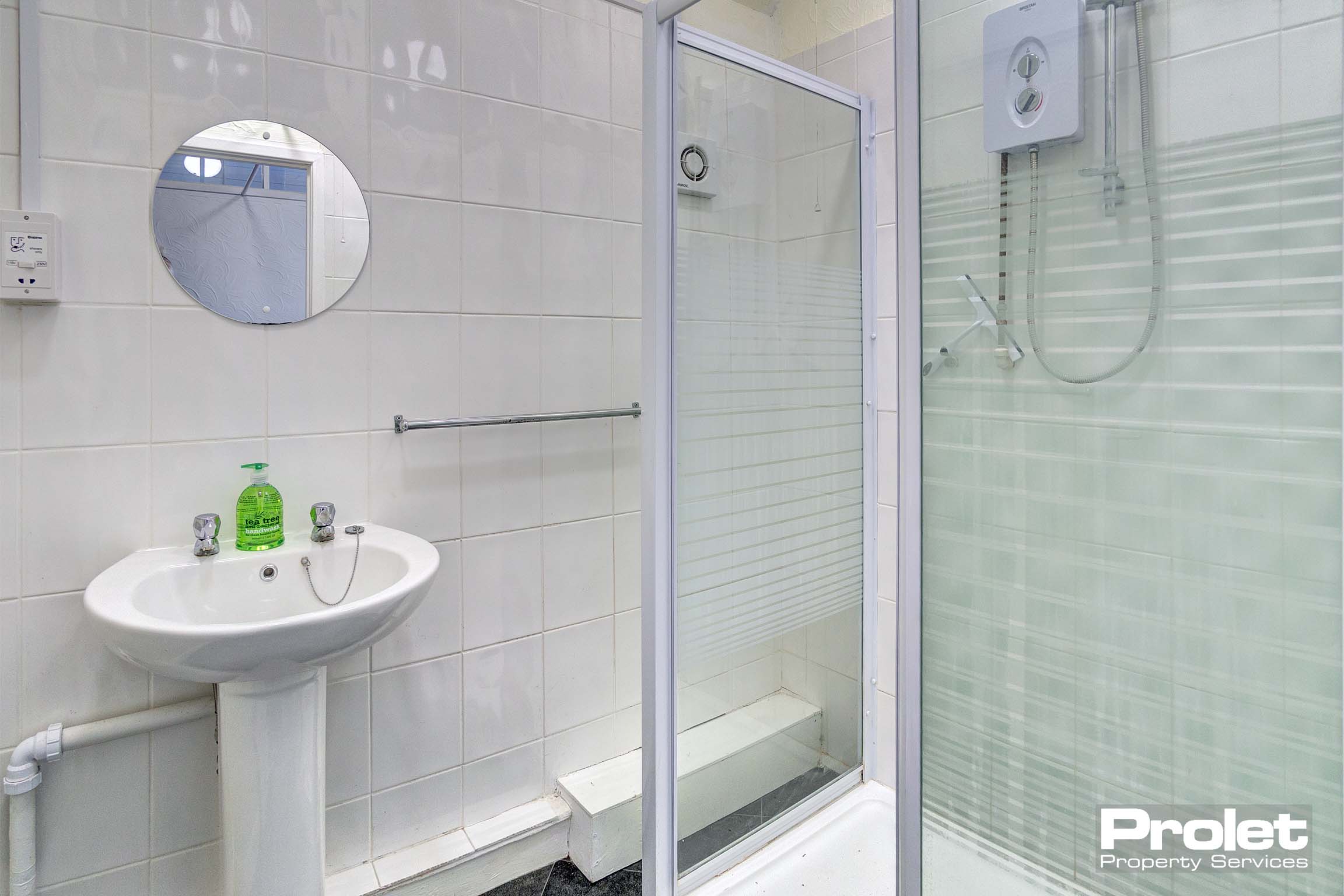 Shower cubicle with electric shower. Grey vinyl floors with white tiled walls and white sink. Mirror attached to wall