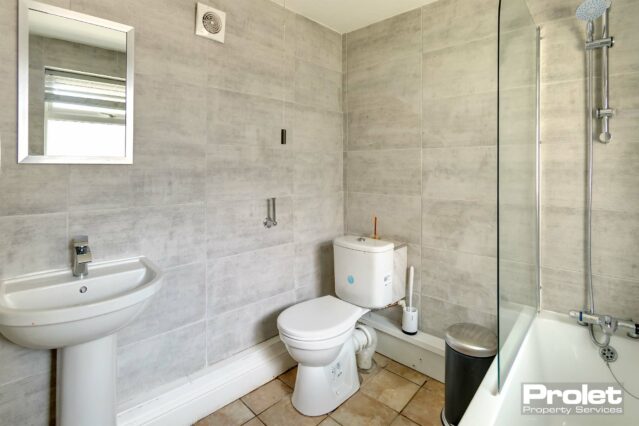 Bathroom off back of kitchen with speckled beige tiled walls with tiled floor. Shower off taps over bath.